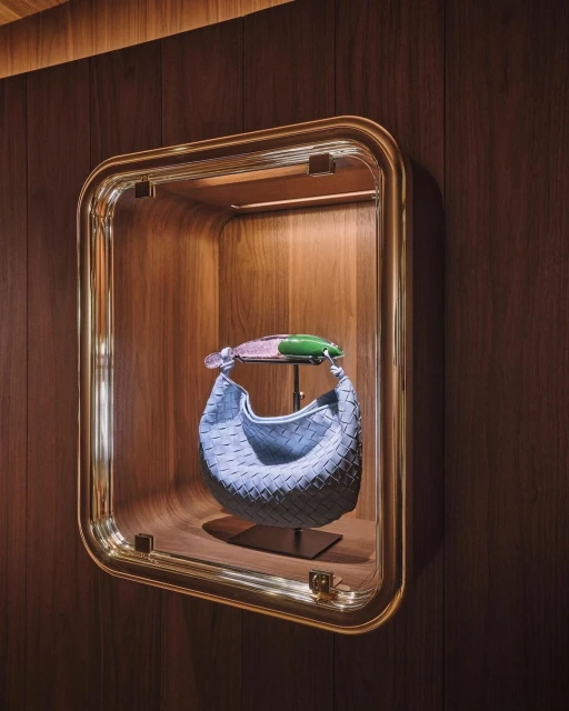 Fashionable blue Bottega Veneta purse on a display stand inside a wall-mounted, gold-trimmed, glass display case with wood panelling.