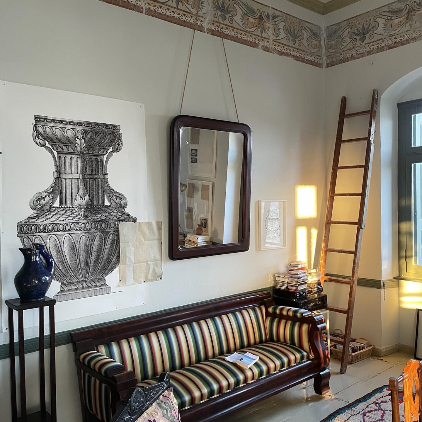 Interior shot featuring a striped sofa, hanging dark wood mirror, wooden ladder, and large architectural vase print on a pale wall.