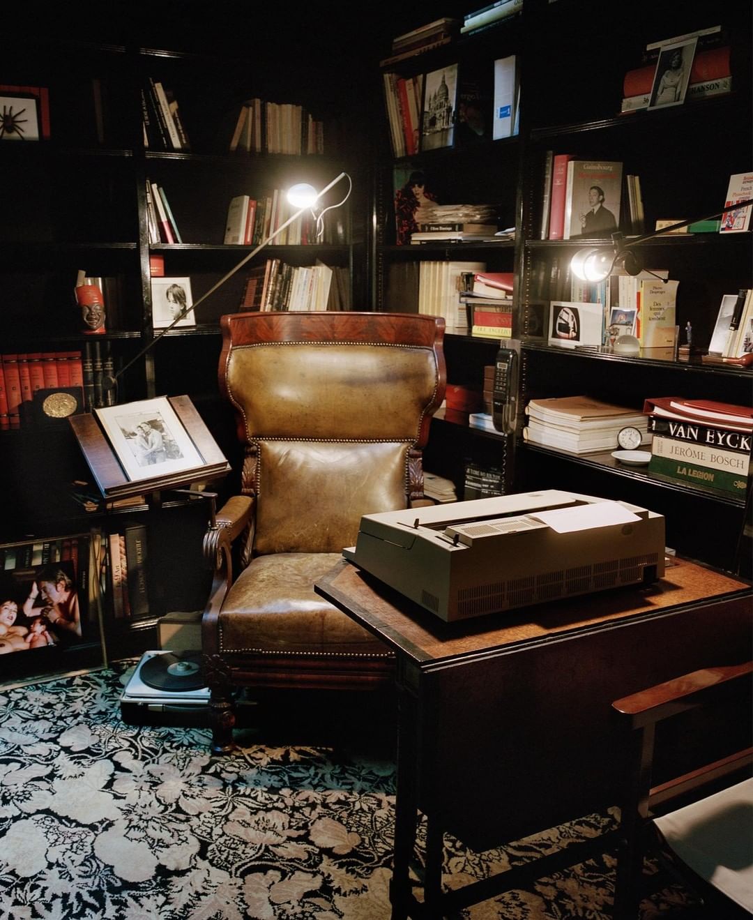 A vintage room features a leather armchair, a side table holding a typewriter, and bookshelves filled with books under warm lighting.