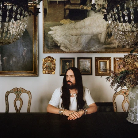 A man with long dark hair and a beard seated at a dark wooden dining table in an ornate room decorated with framed paintings and crystal chandeliers.