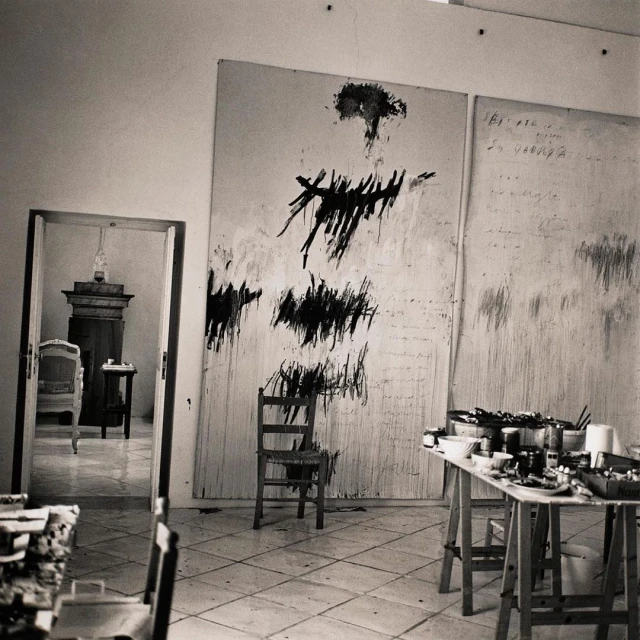 Monochrome photograph inside an artist's studio with painting supplies, a chair and a large black and white abstract painting on a canvas.