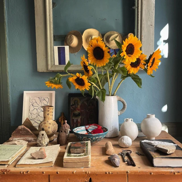 Still life photo of sunflowers in a vase on a wooden table, surrounded by books, pottery, and other decorative objects, with a blue wall.