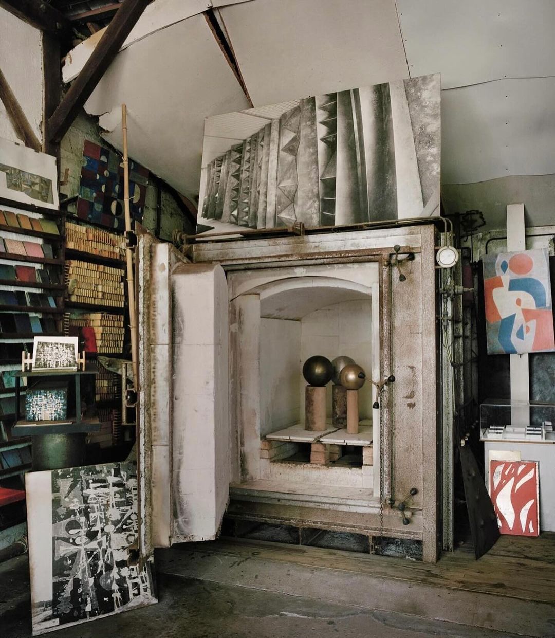 Interior view of an artist's studio with an open kiln showcasing three spherical art pieces, surrounded by artwork and studio equipment.