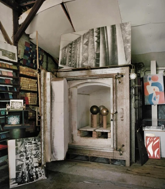 Interior view of an artist's studio with an open kiln showcasing three spherical art pieces, surrounded by artwork and studio equipment.