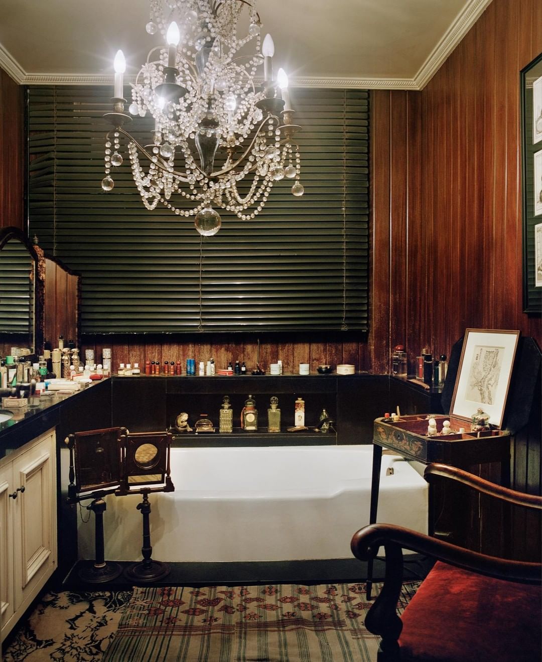 Bathroom interior featuring a chandelier, vanity with perfume bottles, a white bathtub, and wooden wall paneling in a vintage style.