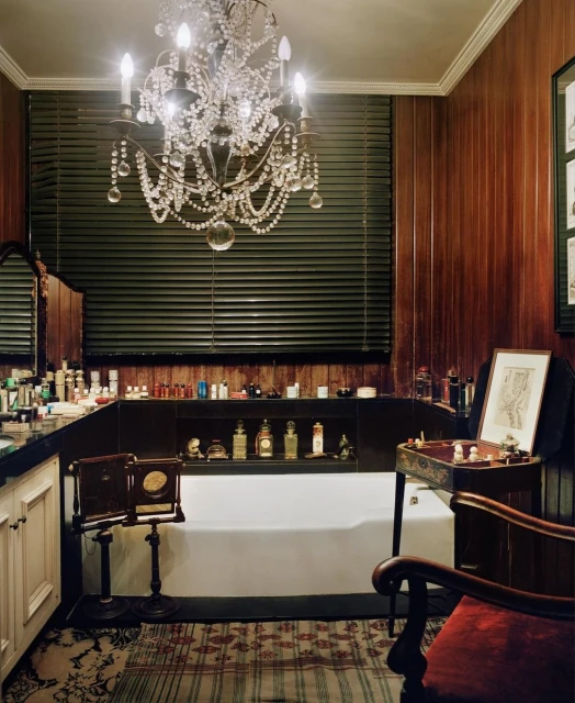 Bathroom interior featuring a chandelier, vanity with perfume bottles, a white bathtub, and wooden wall paneling in a vintage style.