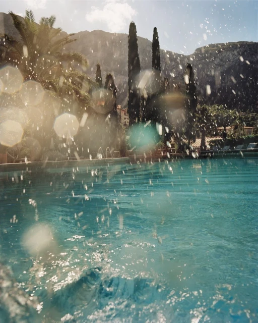 Bright, sunny view of a turquoise swimming pool with splashing water, tall trees, and mountains in the background in a travel setting.