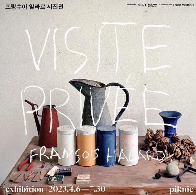 Still life photograph featuring various pitchers, containers, tools, and dry plants arranged on a wooden table against a white wall.