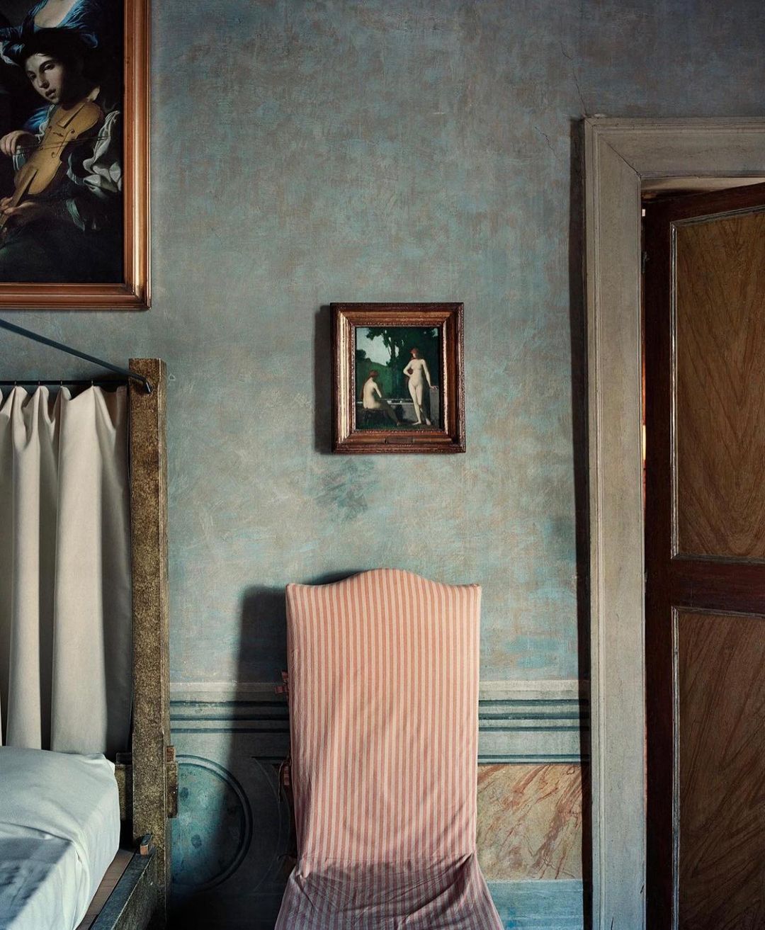 Interior view of a bedroom with a bed, a striped chair, two paintings, and a door, under dim and soft lighting.