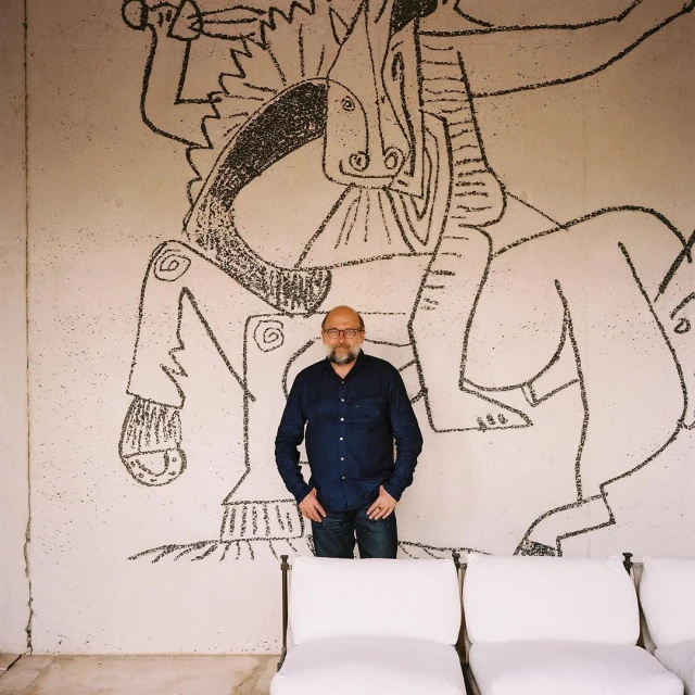 Adult male poses in front of a large wall drawing depicting a person riding a horse, with white chairs in the foreground.