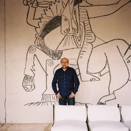 Adult male poses in front of a large wall drawing depicting a person riding a horse, with white chairs in the foreground.