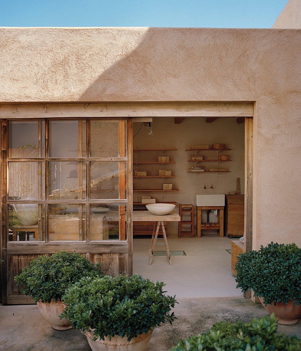 Rustic doorway leads to a minimalist interior with a white ceramic bowl on a trestle table, shelves with objects, and potted plants.