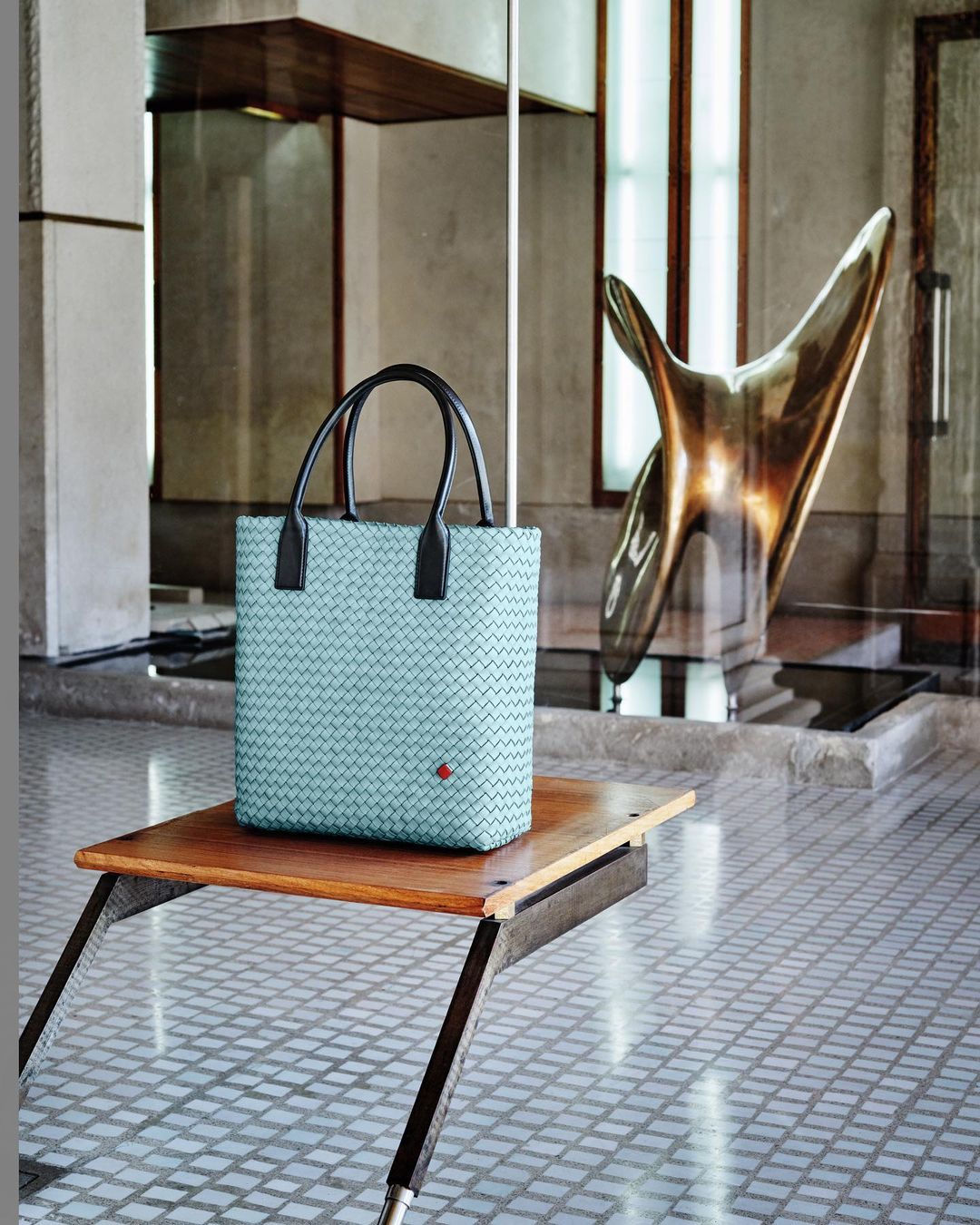 Light blue woven tote bag with black handles, resting on a wooden table against a bright abstract sculpture in an elegant interior.