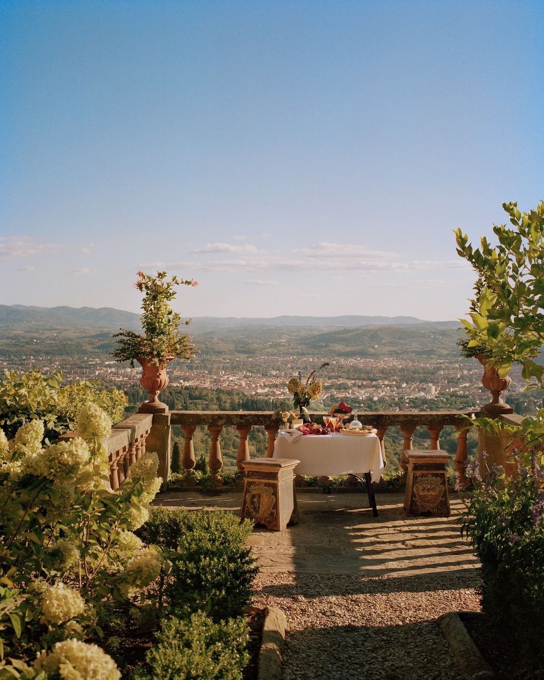 Outdoor terrace with a stone balustrade overlooking a town and distant mountains, set with a table topped with fruit and flowers.
