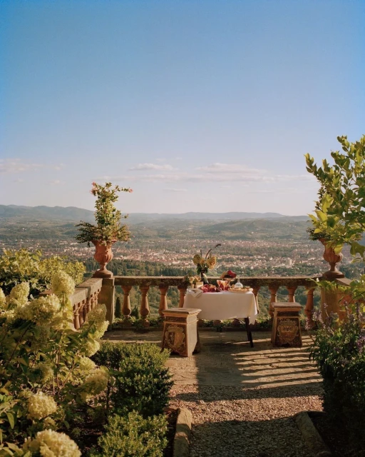 Outdoor terrace with a stone balustrade overlooking a town and distant mountains, set with a table topped with fruit and flowers.
