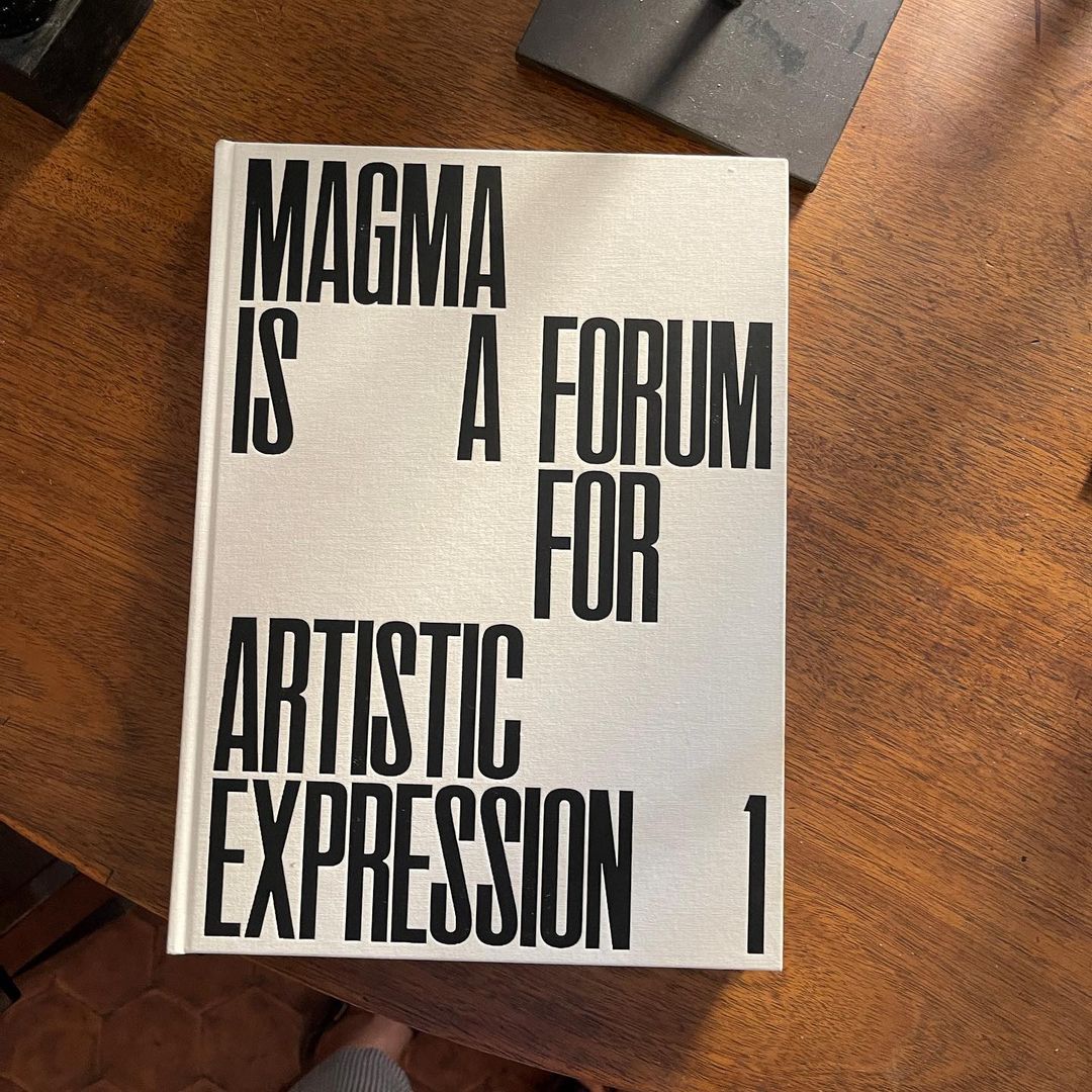 A hardcover book with a white cover and bold black lettering stating 'MAGMA IS A FORUM FOR ARTISTIC EXPRESSION 1' rests on a wooden table.