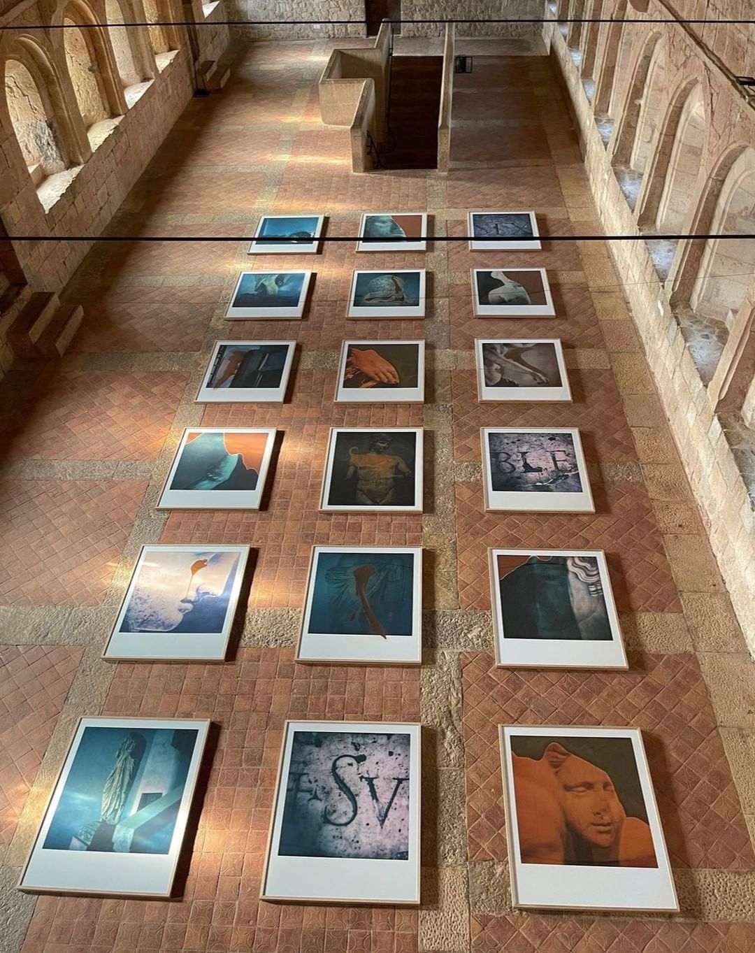 Art gallery exhibition: twenty framed prints arranged in rows on a terracotta-tiled wall, viewed from a high angle, bathed in natural light.
