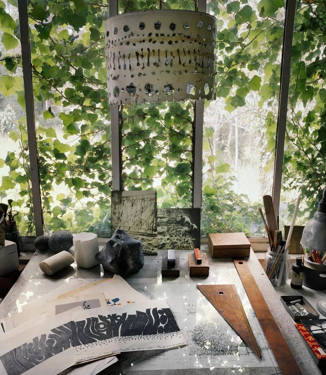Artist's studio featuring a worktable covered in prints, wooden tools, and stones, with natural light streaming through a vine-covered window.