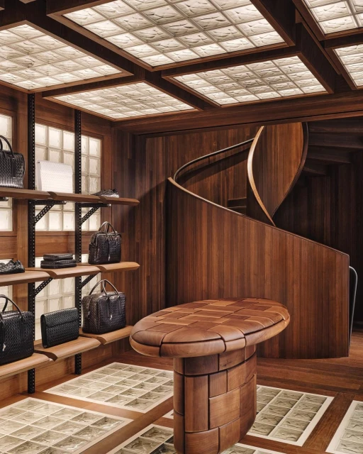 Interior of a retail space featuring shelves with handbags, a wooden staircase, an oval display table, and a ceiling with glass block panels.