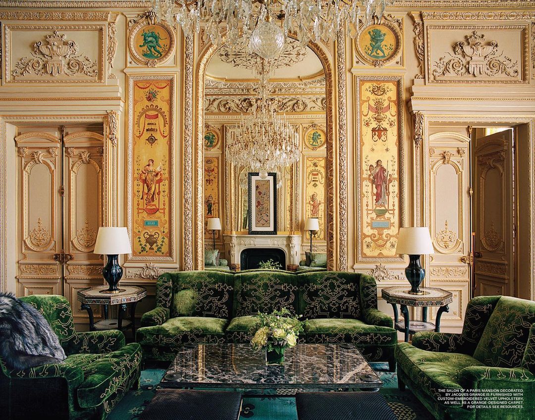 Ornate living room interior with a green velvet sofa and armchairs, tables with lamps, a chandelier, and gold-trimmed architectural details.