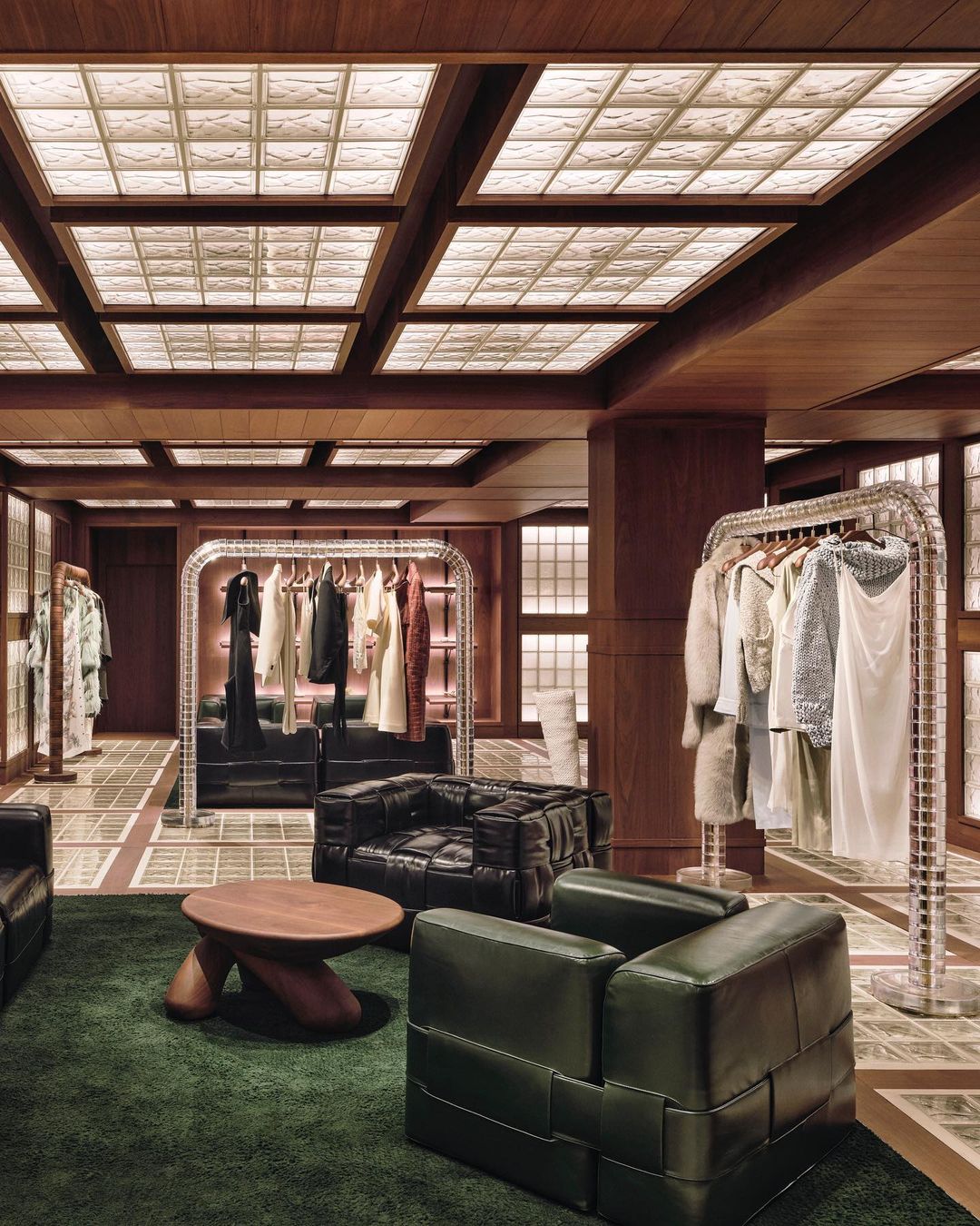 Upscale retail space interior with clothing racks, olive green chairs, a geometric wood ceiling, and soft, diffused lighting.