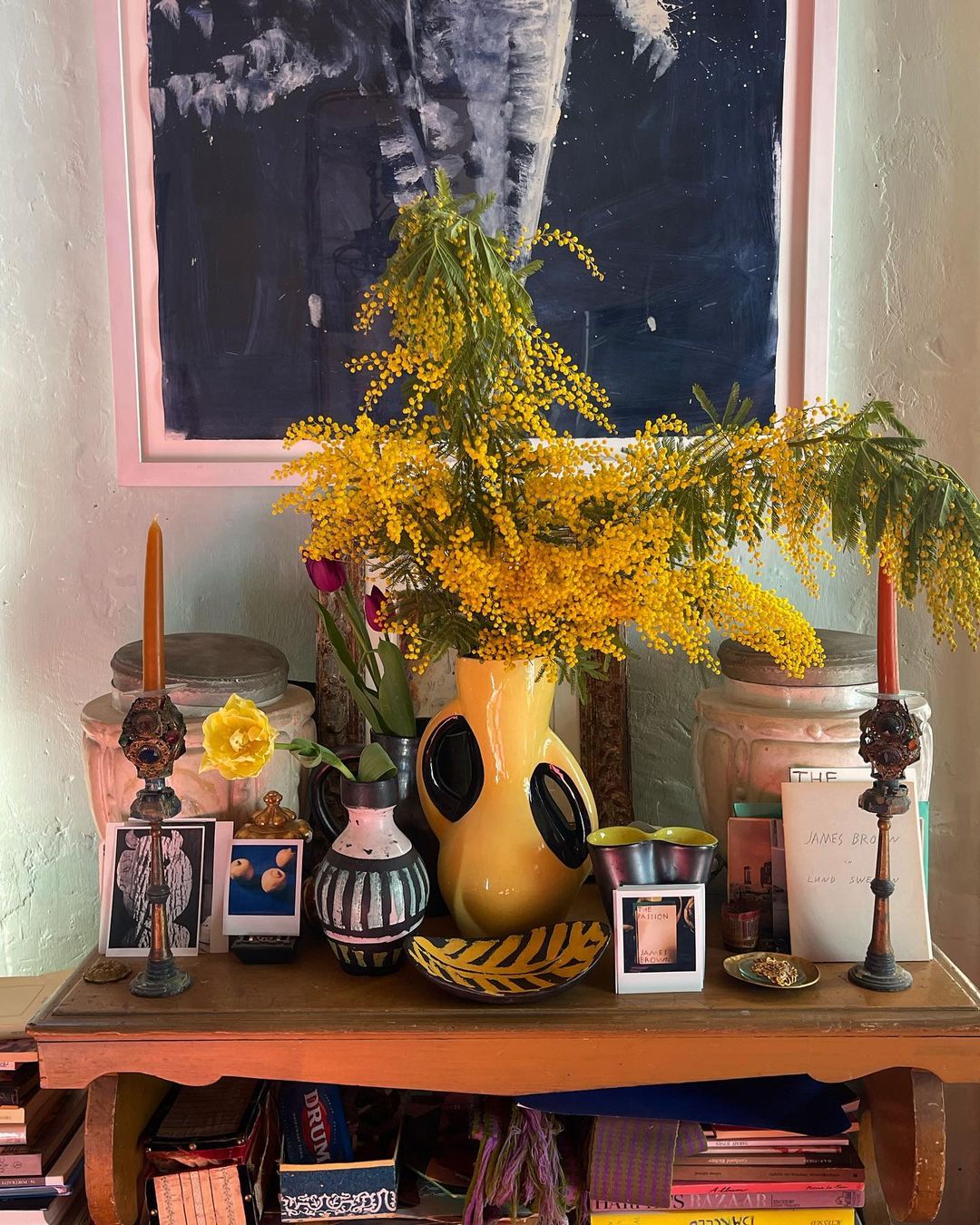 Still life composition featuring vibrant yellow flowers in a vase, alongside antique candlesticks, books, and decorative objects on a table.
