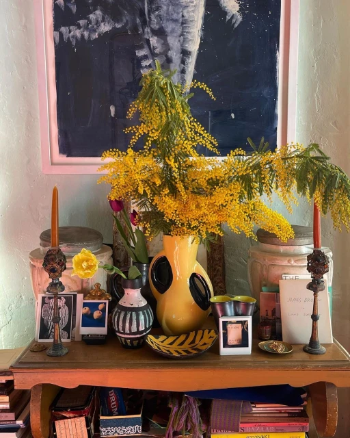 Still life composition featuring vibrant yellow flowers in a vase, alongside antique candlesticks, books, and decorative objects on a table.