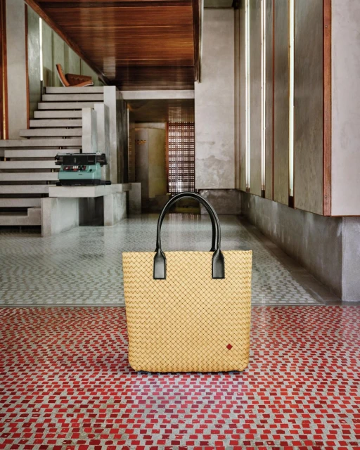 Woven beige handbag with black handles is centered on a red-tiled floor in a modern building with concrete walls and white stairs.