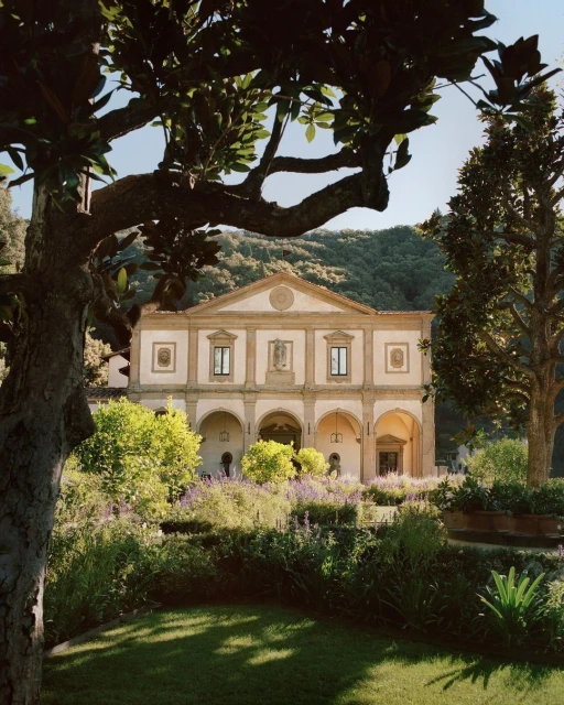 Exterior of ornate Italian villa with arched loggia and lush garden, set against a backdrop of rolling hills, bathed in warm daylight.