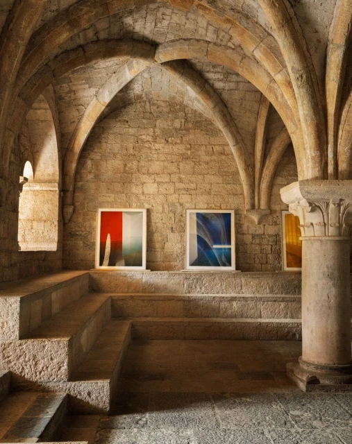 Interior view of a stone building featuring framed abstract art pieces on stepped platforms, illuminated by soft natural light.