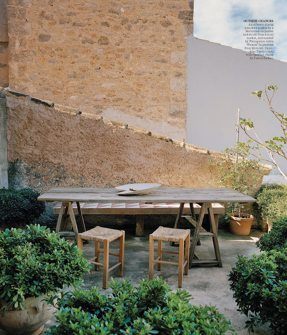 Outdoor dining space with a wooden table, straw stools, and green plants in pots against a stone wall, creating an inviting scene.