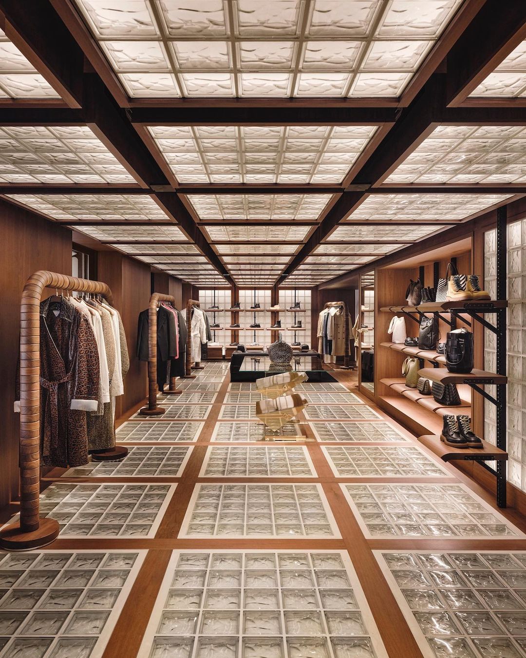 Upscale clothing boutique interior with wood paneling, glass-block floor and ceiling, showcasing garments, handbags, and shoe displays.