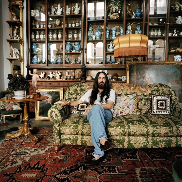 Person sits on an ornate couch in a room filled with decorative objects and furniture, including display cabinets and framed art.