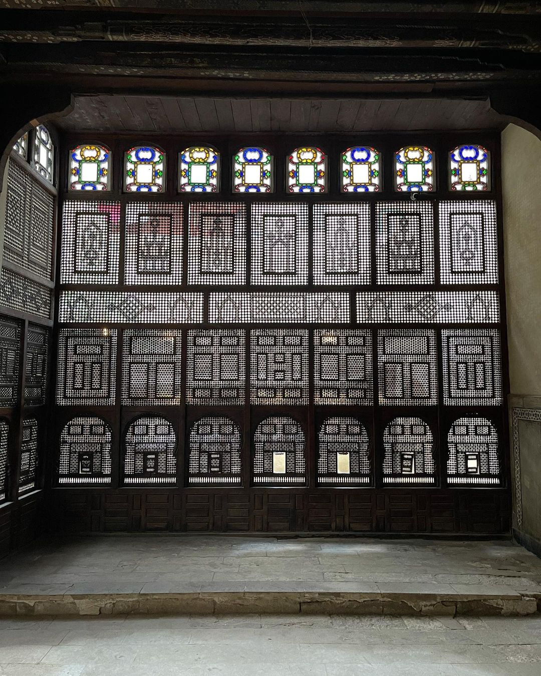 Intricate wooden lattice screen with geometric patterns and stained-glass windows, creating a play of light and shadow in the architecture.