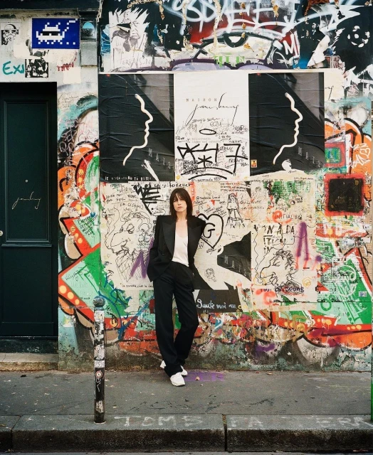 A woman in a suit stands in front of a graffiti-covered wall and posters in an outdoor, urban setting with natural lighting.