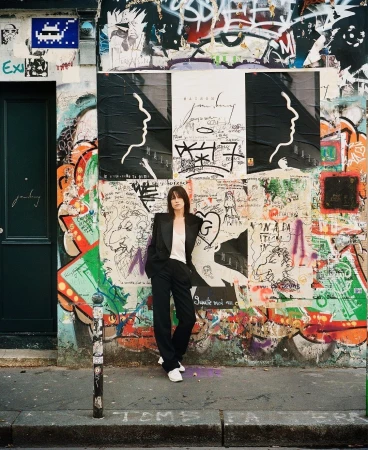 A woman in a suit stands in front of a graffiti-covered wall and posters in an outdoor, urban setting with natural lighting.