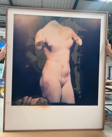 Framed photograph of a classical nude torso in a warehouse setting held up by two people, blending art with industrial space.