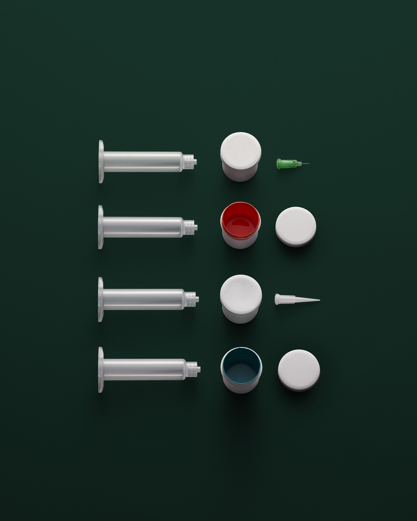 Overhead shot of four syringes, open containers with colorful liquid, and lids on a dark green background, shot in a studio.