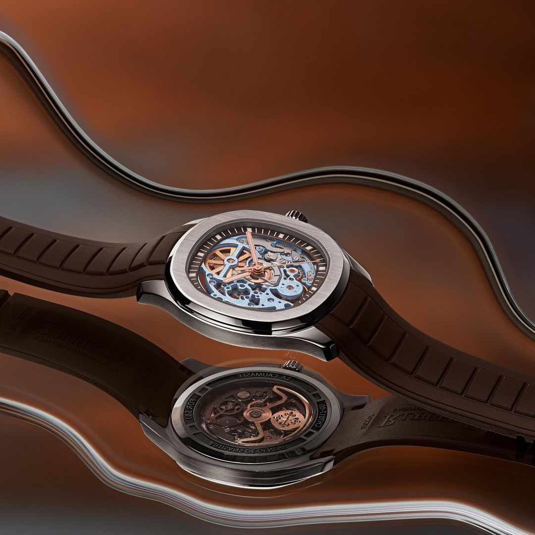 Two luxury wristwatches with a skeleton design are displayed on a reflective brown surface with a metal wire in the background.