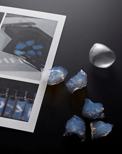 Cosmetic still life: Light blue translucent gel pieces and clear plastic container on a black surface next to a framed photo.