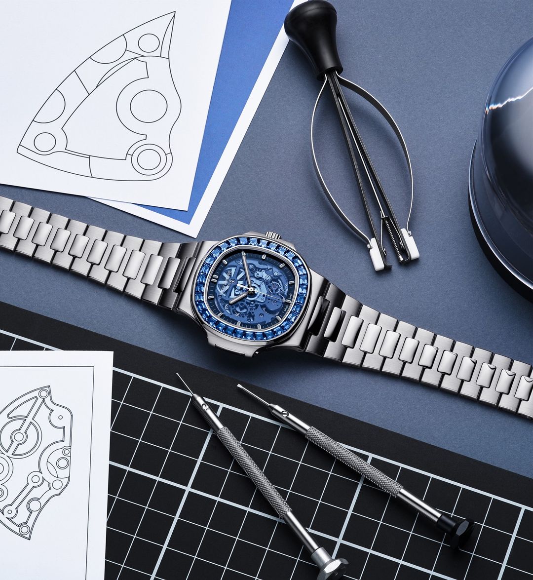 Silver wristwatch with blue face and jewel details displayed with watchmaking tools and technical drawings on a blue surface.