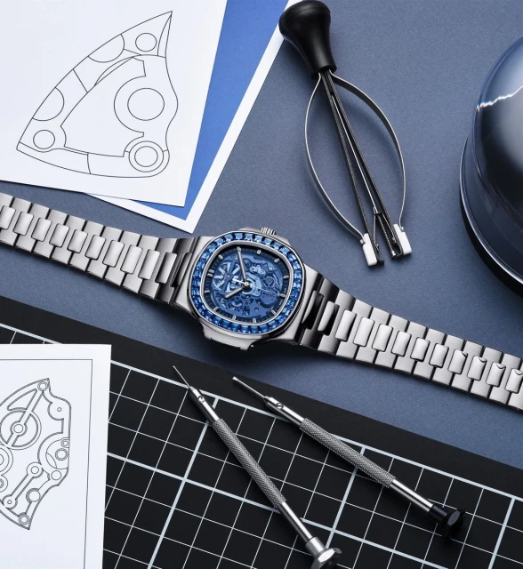 Silver wristwatch with blue face and jewel details displayed with watchmaking tools and technical drawings on a blue surface.