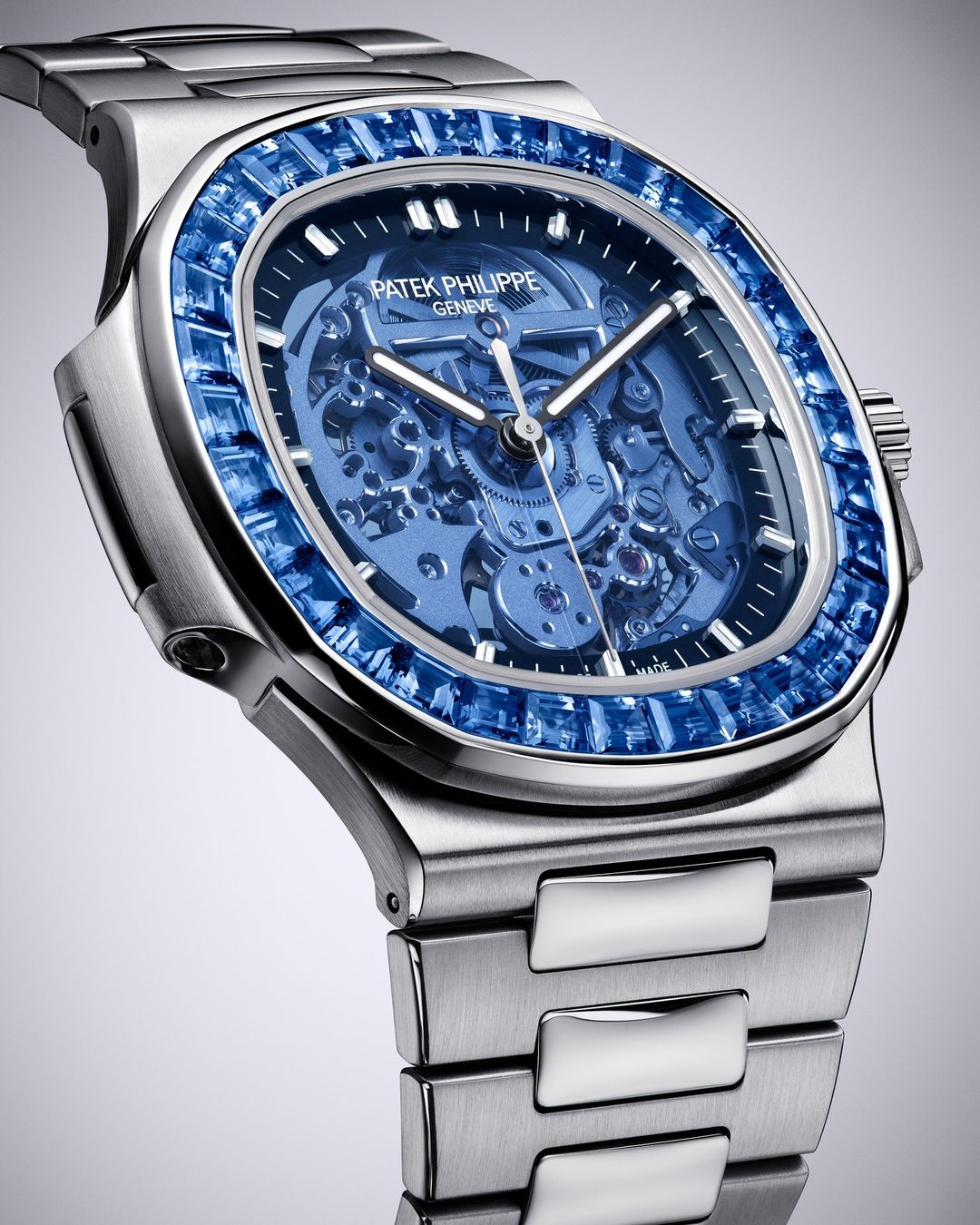 Close-up of a Patek Philippe watch featuring a silver band and a blue sapphire-encrusted bezel against a graduated grey background.
