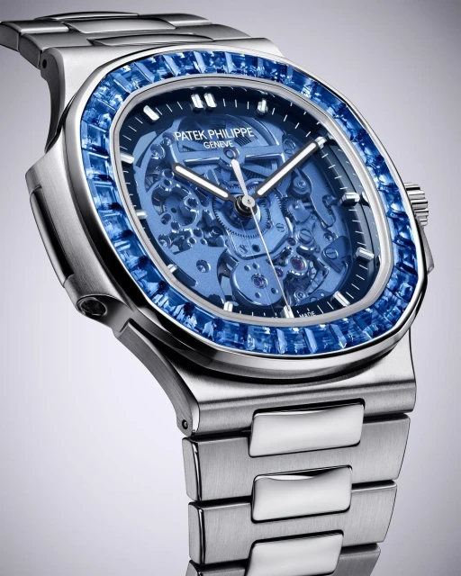 Close-up of a Patek Philippe watch featuring a silver band and a blue sapphire-encrusted bezel against a graduated grey background.
