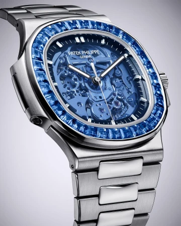Close-up of a Patek Philippe watch featuring a silver band and a blue sapphire-encrusted bezel against a graduated grey background.