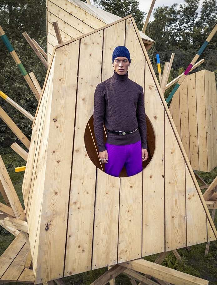 Man in a blue hat and turtleneck stands in a wood structure with purple pants, in a wooded setting, featuring a unique fashion style.