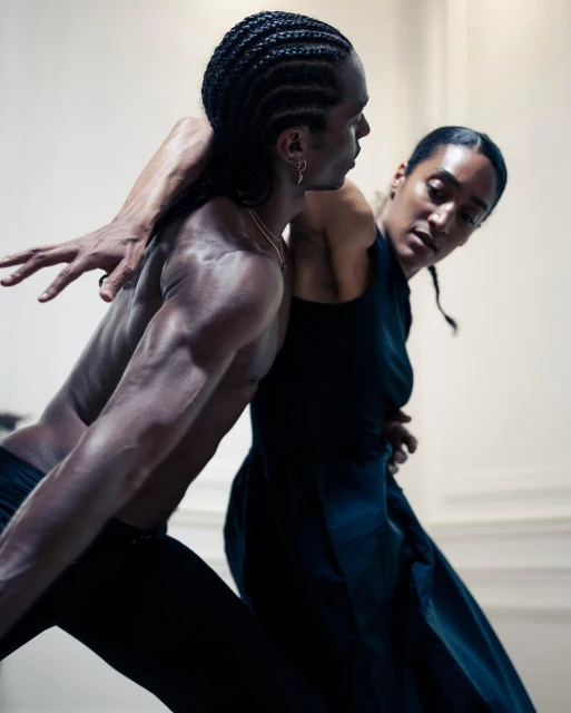 Two athletic dancers, a man and woman, perform a modern dance routine in a bright, minimalist space with dramatic, expressive poses and dark clothing.