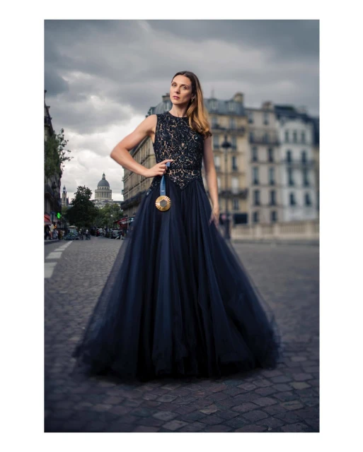 Woman in elegant navy dress with gold medal poses on cobblestone street in Paris, France, with classic buildings and cloudy sky.