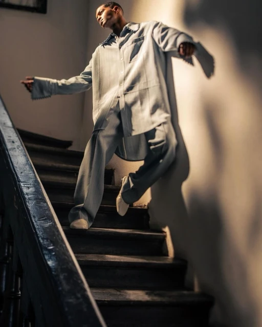 Man in a light blue oversized shirt and pants is going down dark wood stairs in a bright, indoor space with a white wall.