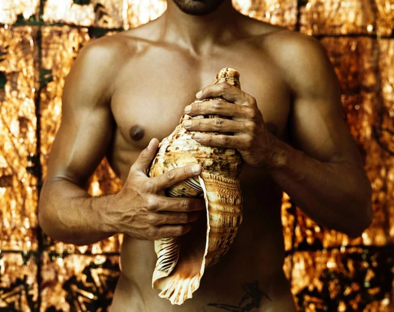 Topless adult male with tattoo, medium shot, holding giant seashell in studio with gold abstract background. Beauty, fashion, body art.